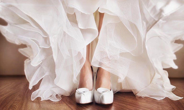 Bride’s white satin shoes peeking from beneath a layered tulle wedding dress on a wooden floor