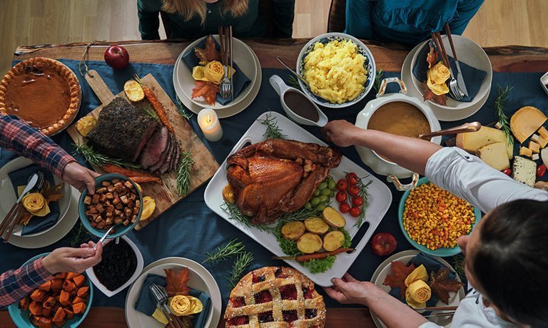 Overhead view of a holiday dinner table with roasted turkey, pies, side dishes and people passing plates