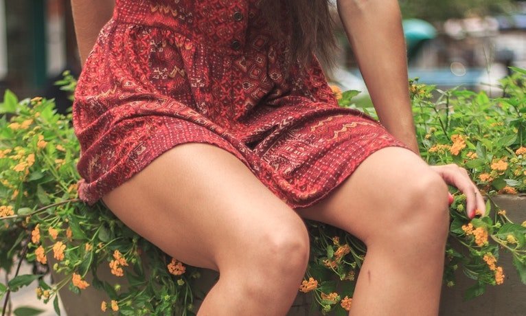 Woman sitting on a planter with knees together wearing a short red patterned dress, showing her inner thighs