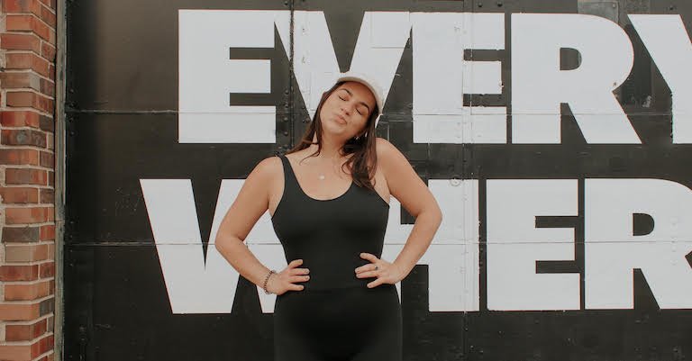 Mid-size model posing with hands on hips in a black fitted sleeveless bodysuit against a black wall with large white letters