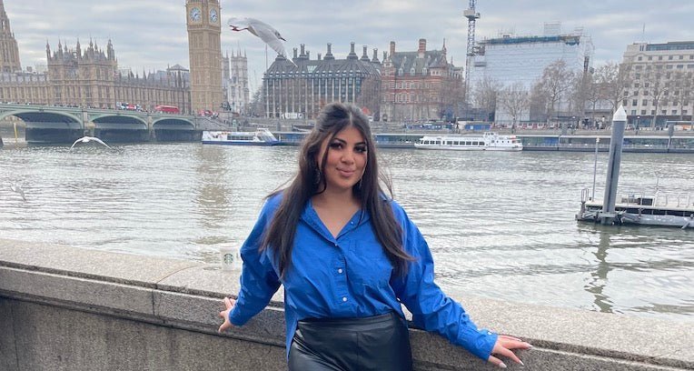 Woman leaning on river railing wearing blue shirt and black skirt with London skyline and Big Ben behind her