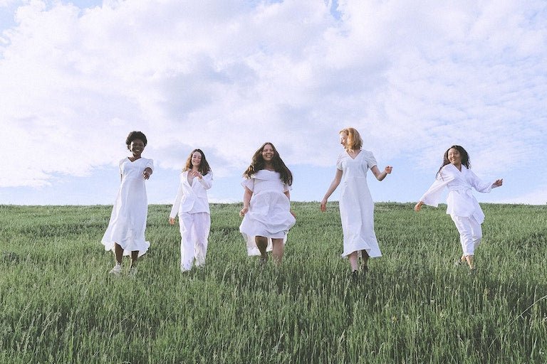Five women walking and laughing across a grassy field wearing flowy white dresses under a cloudy blue sky