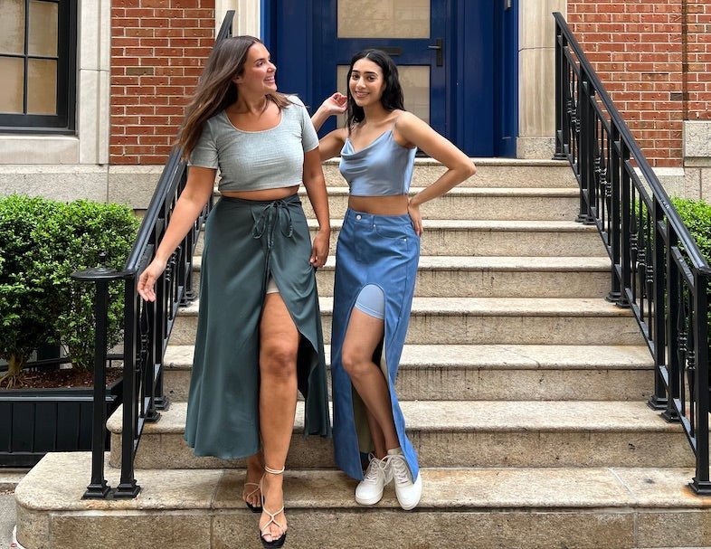 Two women on stone steps, left in sage green tie-waist skirt and cropped tee, right in blue slit skirt and cowl crop top
