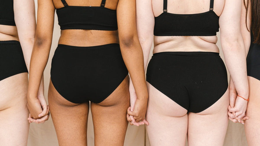 Four diverse women from the back wearing black high rise underwear and bralettes, holding hands against a neutral backdrop