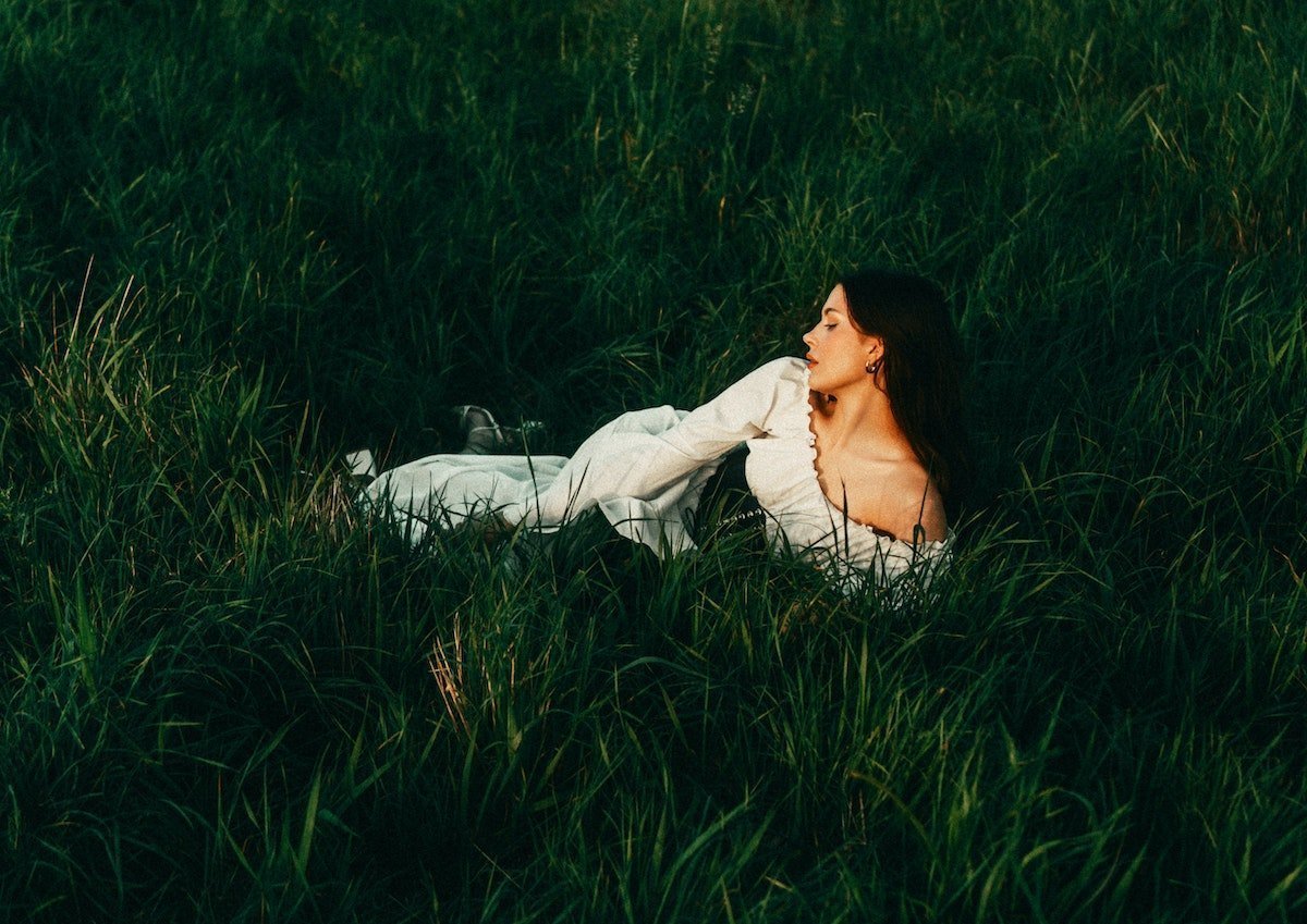Woman reclining in tall grass wearing a white off-the-shoulder, flowy summer dress in a meadow