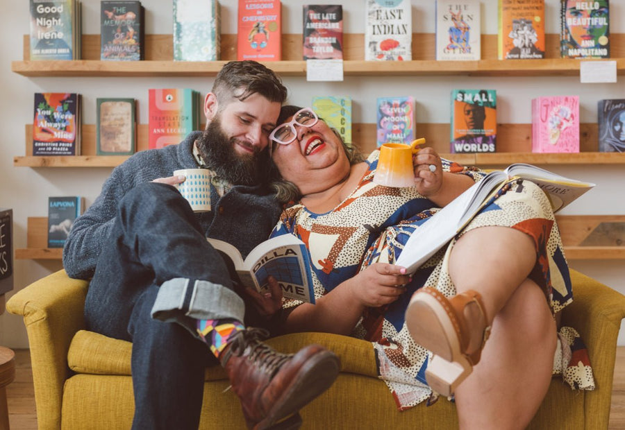 Plus-size bride-to-be laughing with partner on a yellow armchair in a bookstore, holding a mug and a magazine