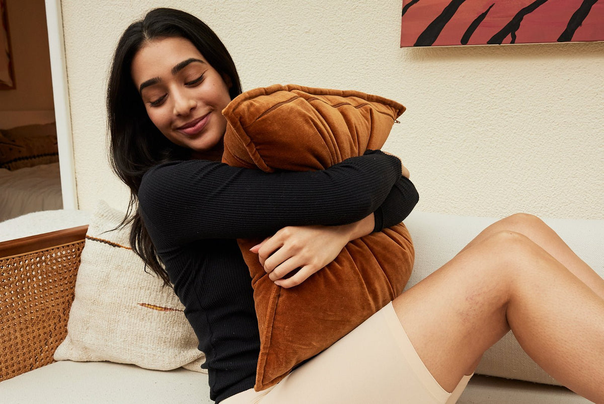Woman seated on a couch hugging a rust-colored velvet pillow, relaxed in a cozy living room setting