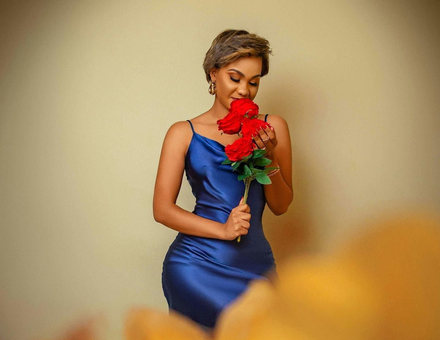 Woman in a fitted blue satin slip dress holding a small bouquet of red roses against a plain beige background