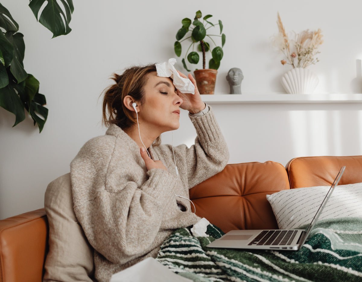 Woman on a couch wiping her forehead during a hot flash, wearing earbuds and a cozy sweater with a laptop on her lap