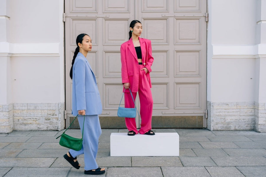 Two models in oversized powder blue and hot pink suits pose with bags before a paneled door, one on a white pedestal