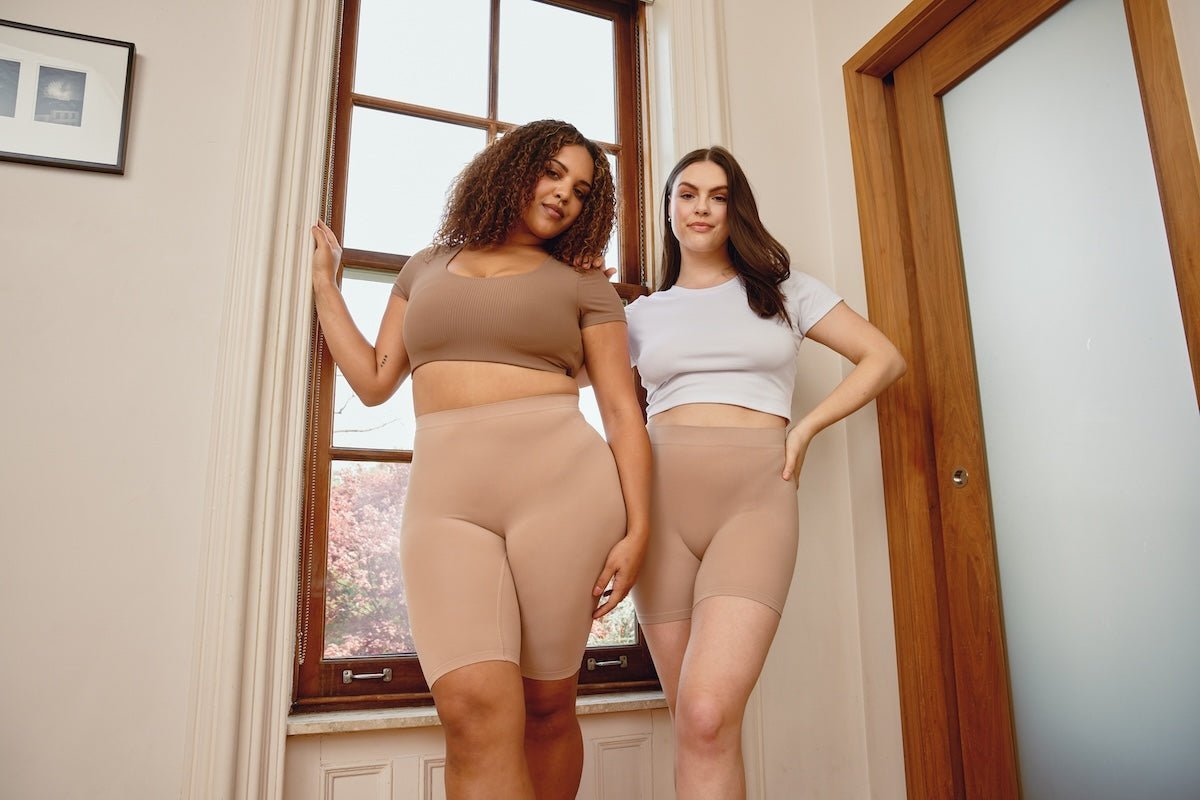 Two models wearing nude no-show high‑waist seamless shaping shorts standing by a sunlit window in a neutral-colored room