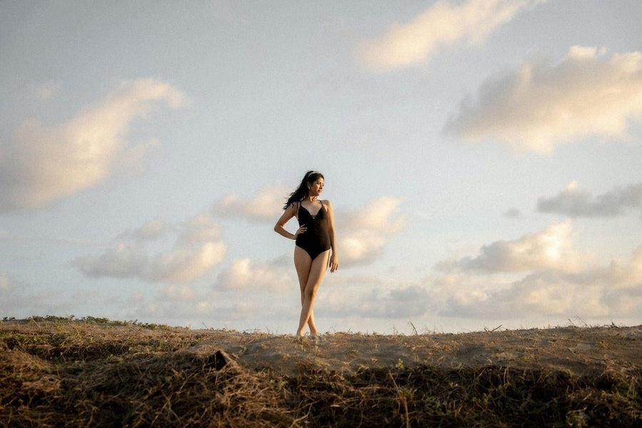 Model wearing a black fitted one-piece bodysuit standing on a grassy hill at golden hour with soft clouds in the sky