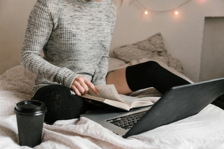Person sitting on a bed reading a book while wearing black thigh-high leg warmers, gray ribbed sweater, with a laptop and takeaway coffee nearby
