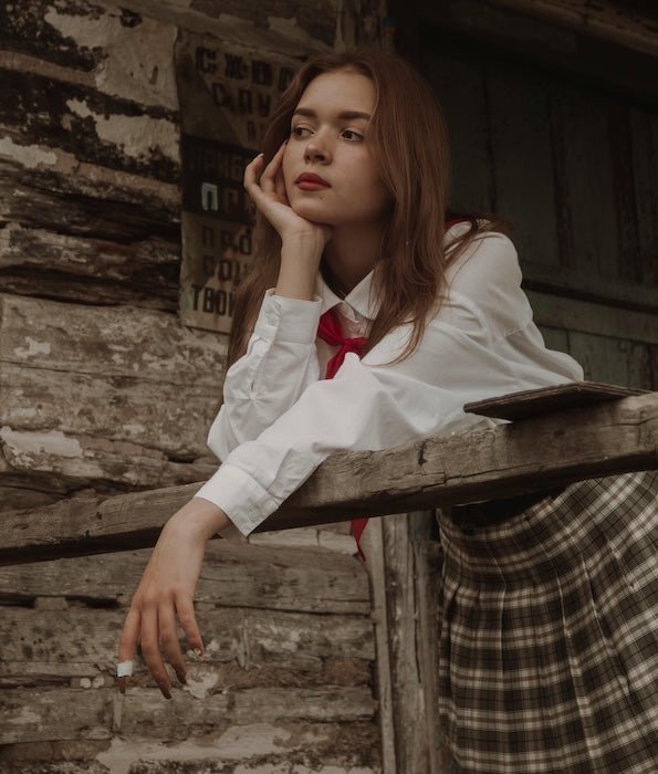 Young woman leaning on a weathered railing, wearing white blouse, red necktie and pleated plaid skirt in a rustic setting