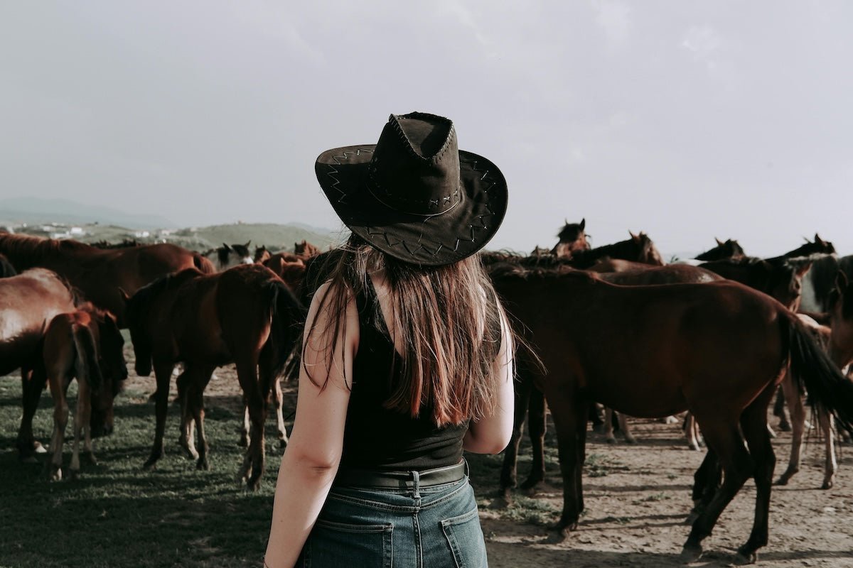 Woman wearing a black cowboy hat, black tank top, and jeans standing with a herd of brown horses in an open field