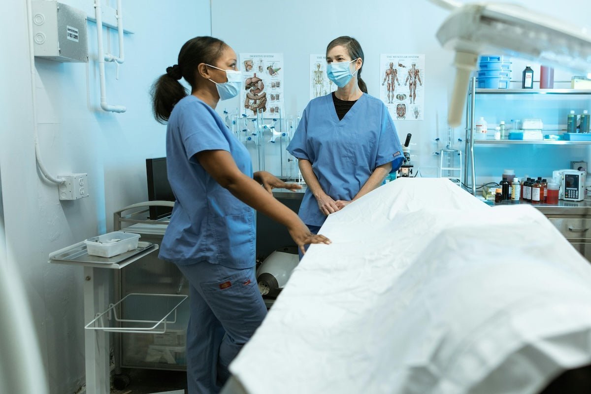 Two masked healthcare workers in blue scrubs talking beside a covered exam table in a clinical room