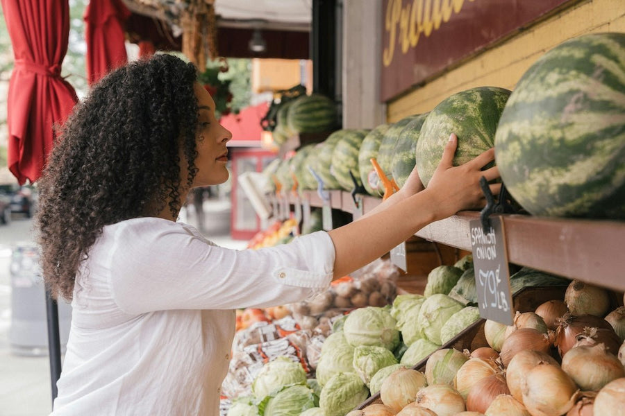 Woman in a white button-down shirt reaching for a watermelon at an outdoor produce stand