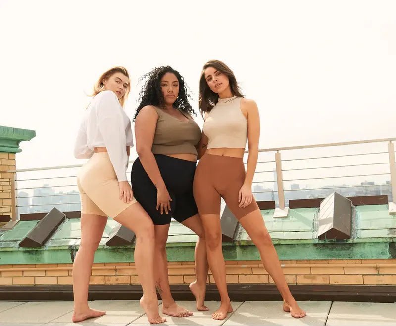 Three women posing on a rooftop wearing high-waist mid-thigh undershorts in nude, black, and brown with city skyline behind